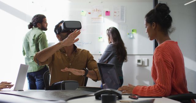 Team of professionals is actively collaborating in an office setting with the use of virtual reality technology. One individual is engaged with a VR headset while others work on laptops. This image can be used to illustrate modern technology in the workplace, innovation, teamwork, and the integration of digital tools in businesses. Suitable for themes on future workplaces, tech advancements, and interactive project development.