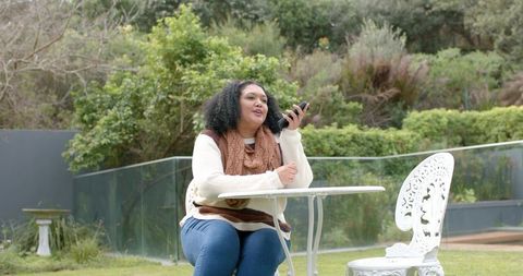 Mid-adult African American woman talking on smartphone at white patio table in garden