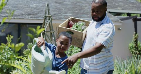 African american father guiding son watering backyard garden and harvesting produce