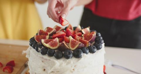 Hands decorating fig and berry layer cake with fresh strawberries, figs and blueberries