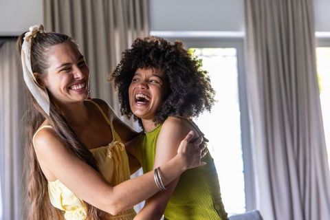 Multiracial women laughing and embracing in sunlit living room wearing yellow and green