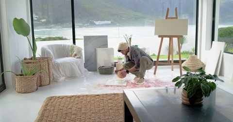 Indian artist crouching and mixing paint on canvas in sunlit coastal studio with easel