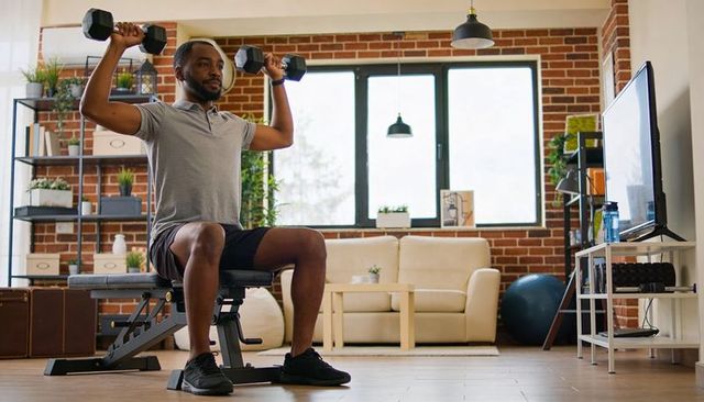 Man performing seated shoulder press with dumbbells on bench in modern home gym living room