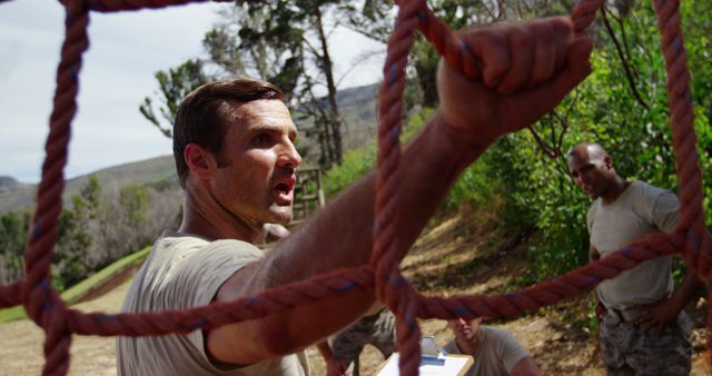 Caucasian man climbs a rope at an outdoor training course. His determination is evident as an African American man observes in the background.