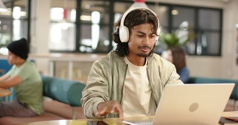 Indian man wearing white headphones using silver laptop in modern coworking lounge