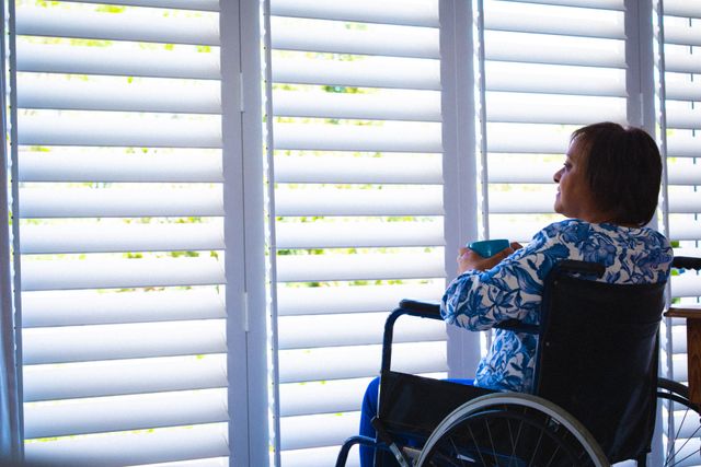 Disabled african american senior woman looking through window shutter while sitting on wheelchair. unaltered, physical impairment, retirement and disability concept.