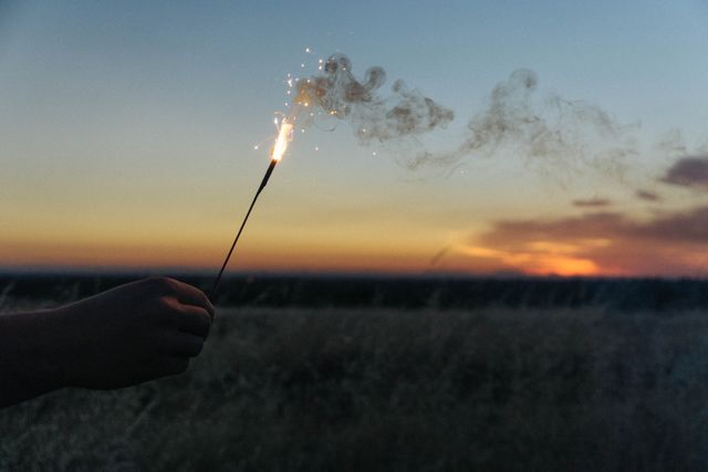 Hand holding sparkler outdoors at sunset in a field with a beautiful evening sky. Great for concepts of celebrations, festivals, summer fun, or social media banners.