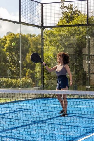 Young woman playing padel on bright blue court with glass walls, serving green ball outdoors
