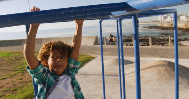 A young boy with curly hair is playing on blue monkey bars at a coastal playground. He is enjoying a sunny day, wearing casual clothing, and displaying excitement. Ideal for promoting outdoor activities, childhood development, family outings, and holiday destinations.