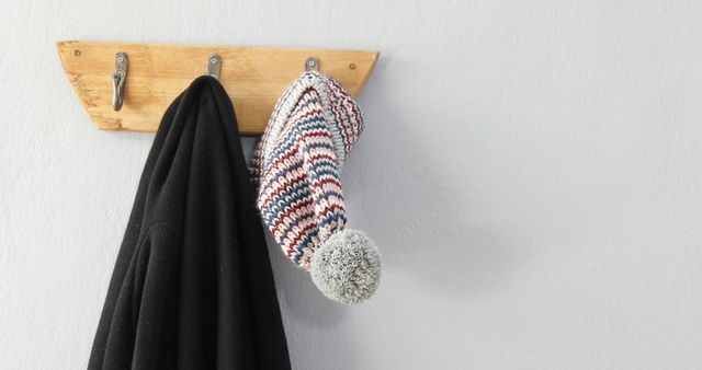 A wooden coat rack mounted on a white wall holds a black coat and a colorful knitted hat with a pom-pom. The simplicity of the scene suggests a tidy and minimalist approach to home organization.
