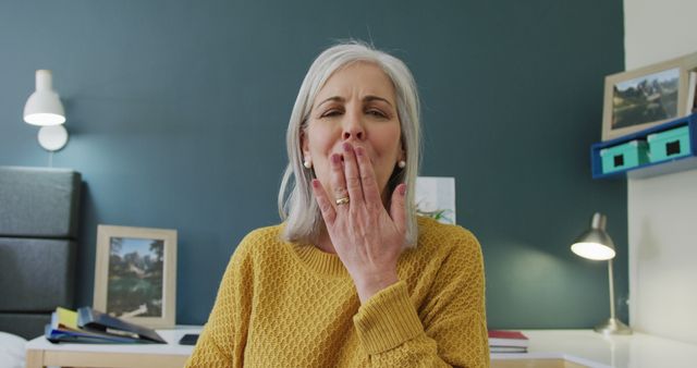 An elderly woman is yawning and looking tired while sitting in a well-decorated room with pictures and warm lighting. This image is perfect for articles or advertisements related to sleep health, relaxation, home comfort, or products for seniors.