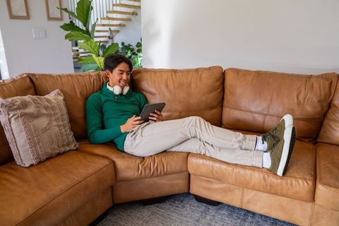Relaxing asian man reclining on tan leather sectional sofa using tablet at home