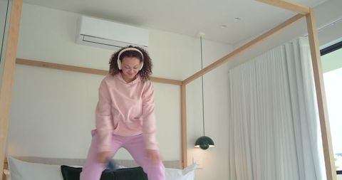 Young african american woman jumping on canopy bed wearing pink hoodie and headphones