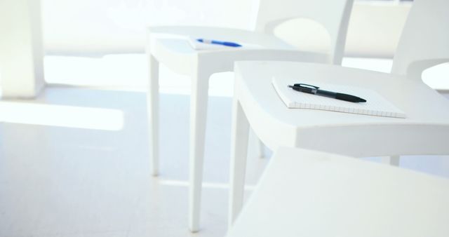 Multiple empty white chairs arranged in a bright room with notepads and pens placed on each, ideal for depicting educational settings, workshops, or business meetings focused on preparation and learning environments.
