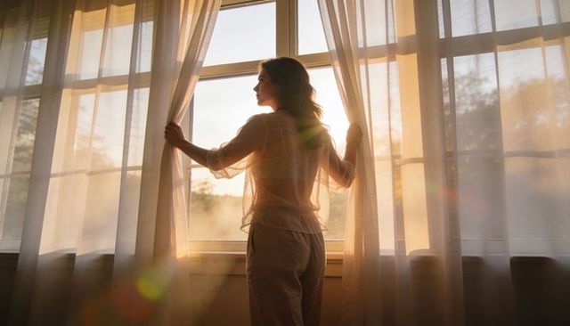 Woman pulling curtains at sunrise silhouette bathed in golden morning light for calm reflection