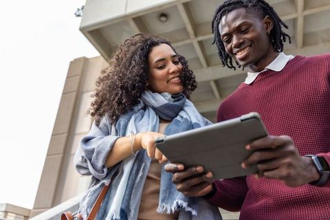 Diverse students collaborating on tablet outside modern campus building, smiling and pointing