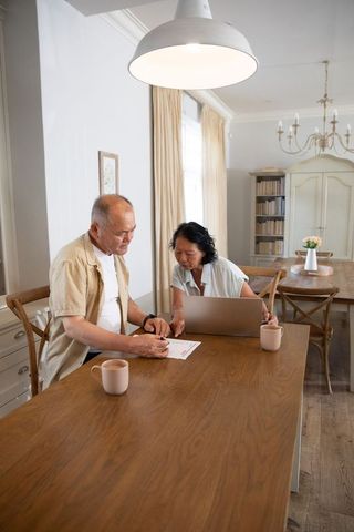 Senior chinese couple reviewing finances and working on laptop at sunlit dining table