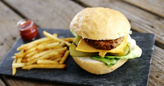 A cheeseburger with lettuce and a side of fries is presented on a slate board, with ketchup in the background. The meal is set on a rustic wooden table, evoking a casual dining atmosphere.