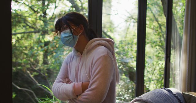 Woman wearing a face mask holding a coffee cup while looking outside through large windows in a home filled with natural light. Ideal for themes of health safety during pandemic, staying safe at home, thoughtful contemplation, or enjoying a peaceful morning indoors.