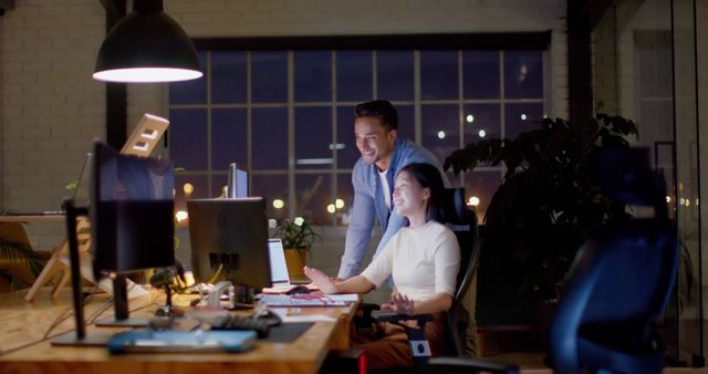 Colleagues engaged in a late night collaboration in a modern office setting. Both are focused and enthusiastic while working on their computers. Ideal for use in business and technology articles, corporate culture promotions, and teamwork concepts.