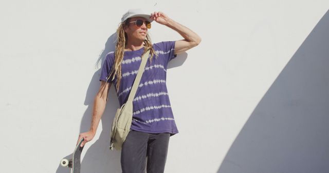 Image of caucasian man with dreadlocks holding skateboard leaning against wall in the sun. Freedom, inclusivity, sport, hobbies and outdoor lifestyle concept digitally generated image.