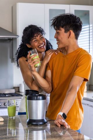 Diverse couple making green smoothies in modern kitchen, laughing and sharing morning warmth