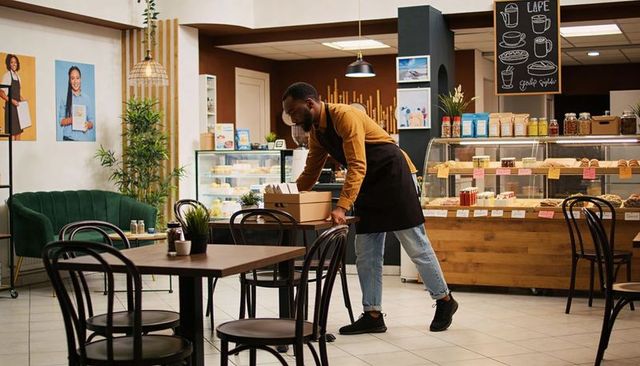 Barista arranging takeout boxes in cozy modern cafe with pastry display and velvet seating
