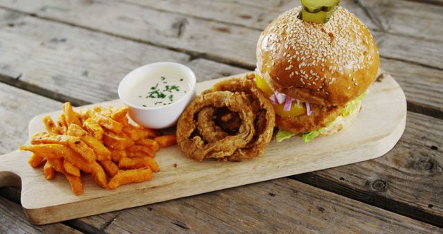 A delicious burger with sesame seeds, crispy onion rings, and seasoned sweet potato fries served on a wooden board, with a side of dipping sauce. This appetizing meal is a classic example of American fast food cuisine, perfect for those craving a hearty and indulgent dish.