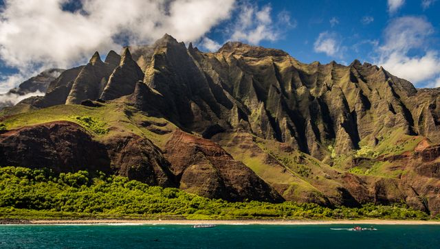 Beautiful view of Na Pali Coast in Kauai showcasing stunning mountain peaks and lush greenery with azure ocean and blue sky in background. Ideal for use in travel brochures, nature magazines, photography collections, and promotional materials about Hawaii or natural landscapes.