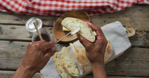 Spreading butter on rustic ciabatta slices at wooden picnic table with jam and checkered cloth