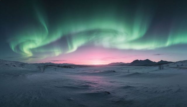 Aurora borealis swirling over snowy arctic tundra at pink dawn with mountain silhouettes