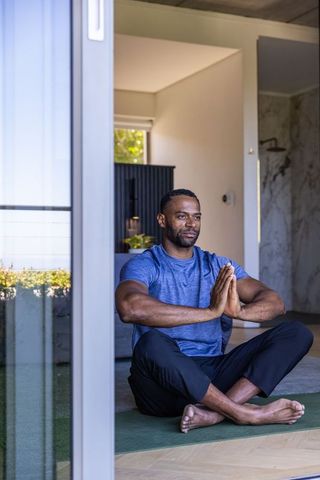 Mature man meditating on yoga mat by sliding glass door in minimalist home with sunlight
