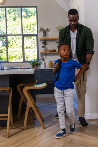 African american father helping son adjust backpack in modern kitchen morning routine