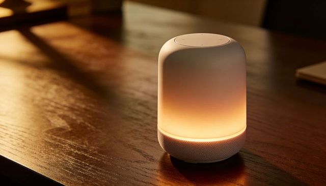 Smart speaker glowing amber casting warm ambient light on polished wood table with mesh band