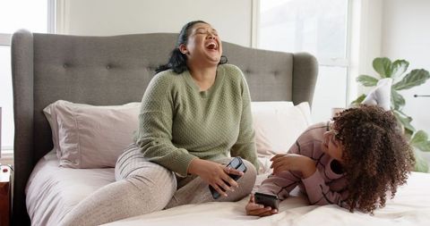 Mother and Daughter Laughing on Sunlit Cozy Bed Holding Mug and Phone