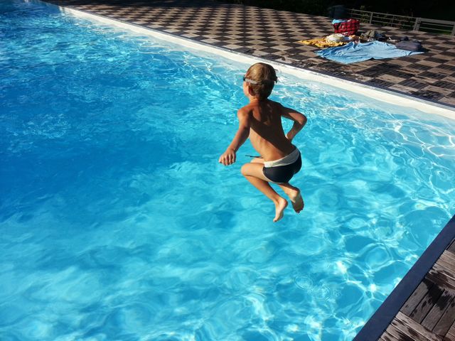 Young boy in mid-air jumping into bright blue pool water. Ideal for themes of summer fun, outdoor activities, swimming, and childhood joy.