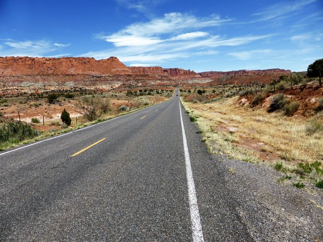 This image captures a vast desert highway running through an iconic red rock landscape under a vibrant clear sky. Ideal for use in travel blogs, adventure advertisements, and scenic backdrop features. It portrays themes of freedom, solitude, and the expansive beauty of the American Southwest.