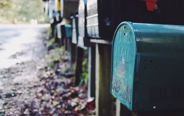 This image shows a row of rural mailboxes along a country road, sun reflecting on weathered surfaces. Perfect for illustrating rural lifestyle, postal delivery routes, community amenities, countryside settings or mail services topics.