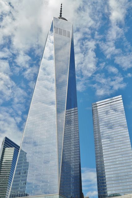 Impressive view of One World Trade Center in New York City with clear blue sky and scattered clouds reflecting on its glass surface. Useful for articles on modern architecture, business environments, urban landscapes, and travel destinations.