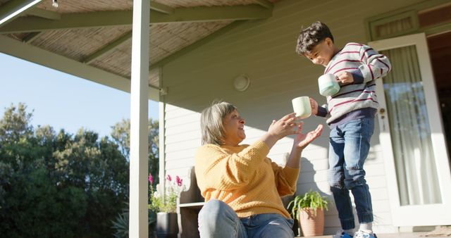 Grandmother seated on porch steps, enjoying tea with grandson standing nearby. They are both holding cups and sharing a joyful moment on a sunny day. Ideal for themes of family bonding, intergenerational relationships, leisure activities, and outdoor relaxation.