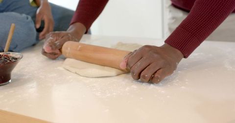 Hands of diverse couple rolling dough on kitchen counter with wooden rolling pin and jam