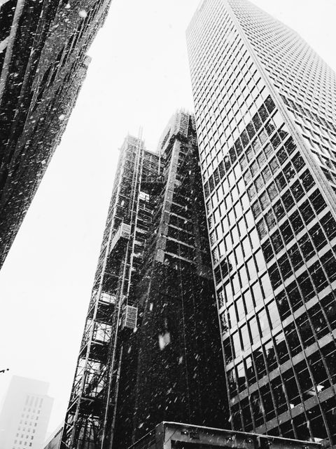 Snow falls on modern skyscrapers in an urban downtown area. Tall buildings create a dramatic cityscape under winter conditions. Use this image to depict cold weather in an urban environment, city life, or for architectural and construction-themed concepts.