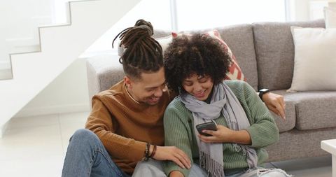 African american couple relaxing on floor using smartphone in bright modern living room