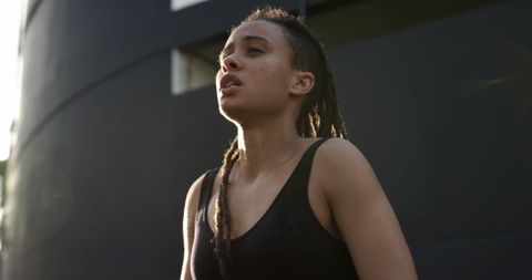 Young African American woman with locs captured in an urban environment, suggesting an active and confident lifestyle. She exudes determination while lit by the evening sun. Perfect for promoting health, fitness, and positivity. Useful for advertisements focusing on city living, women's empowerment, or athletic brands.