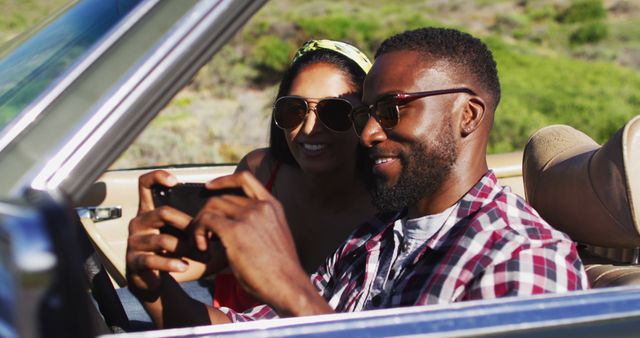 African american couple using smartphone while sitting in the convertible car on road. road trip travel and adventure concept