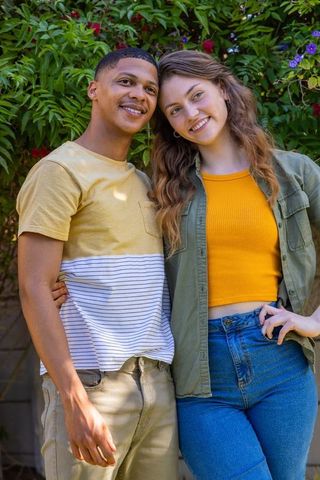 Smiling diverse young couple posing in sunlit backyard garden with blooming shrubs