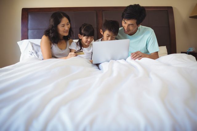 Family sitting on bed using laptop, enjoying quality time together. Suitable for themes of family bonding, technology use at home, online shopping, and modern parenting. Ideal for advertisements, blogs, and articles about family life, technology, and home comfort.