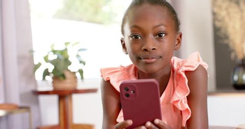 African girl holding smartphone and looking at camera, wearing pink ruffled top at home