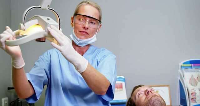 Female dentist in blue scrubs adjusting a dental light while examining a patient in a clinic. Useful for articles and materials on dental health, medical professions, patient care, and dentist-related services.