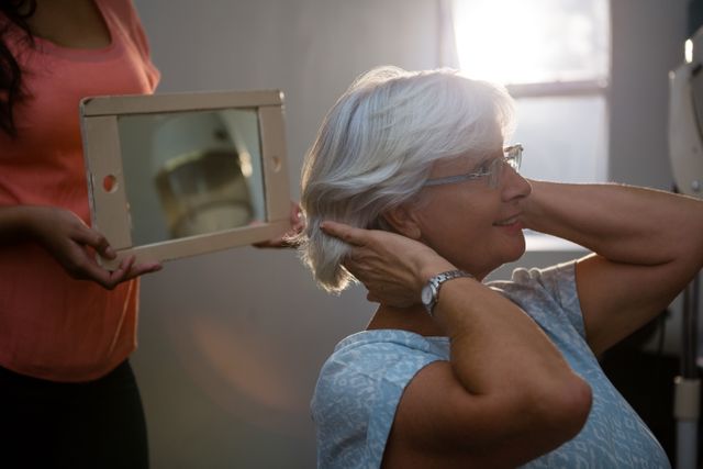 Senior woman admiring her new hairstyle in a salon, with a hairstylist holding a mirror. Ideal for use in advertisements for beauty salons, hair care products, and services targeting elderly clients. Can also be used in articles about self-care and grooming for seniors.